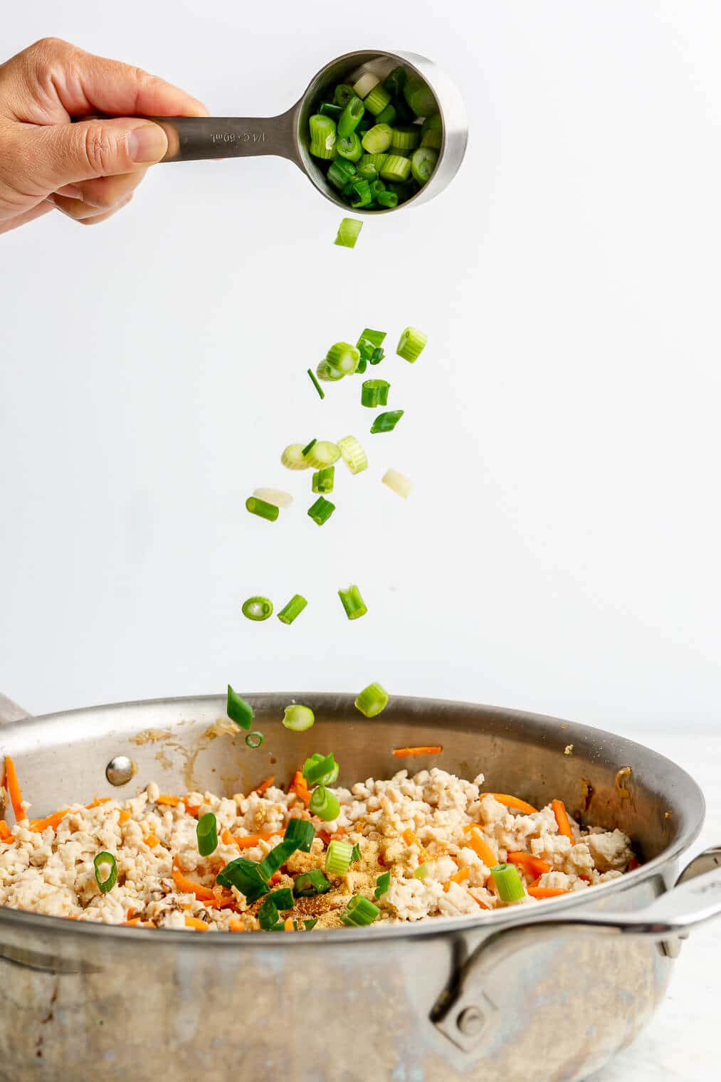 A person adding diced green onions to a skillet of ground turkey and shredded carrots.