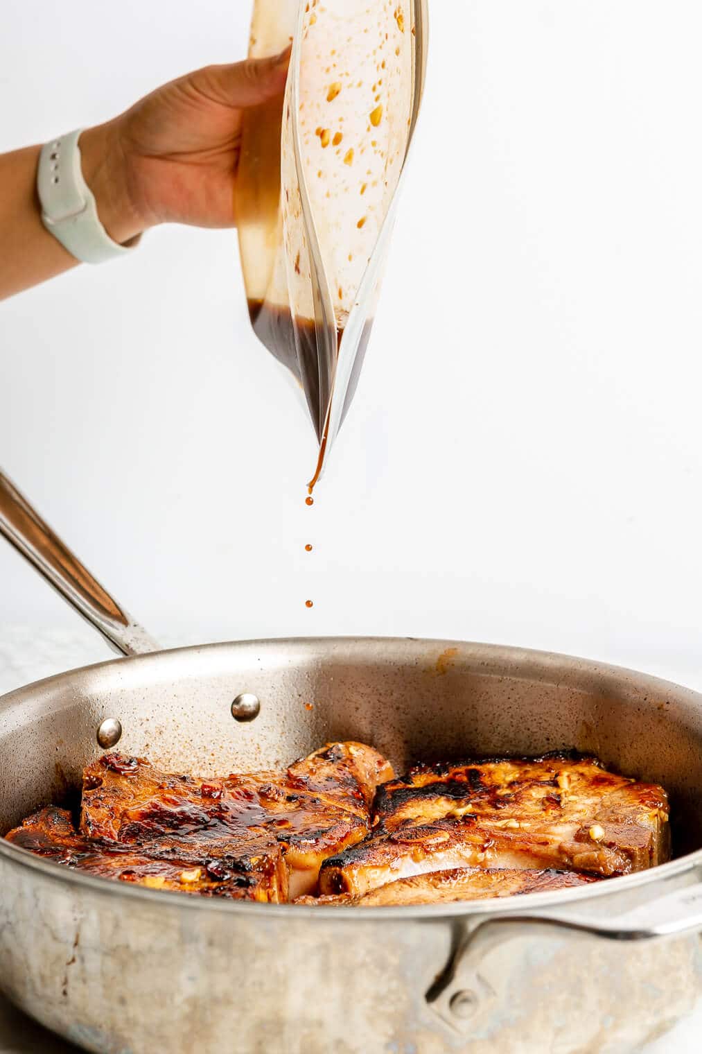 A person pouring a honey garlic pork chop marinade over top pork chops in a large high-walled stainless steel skillet.