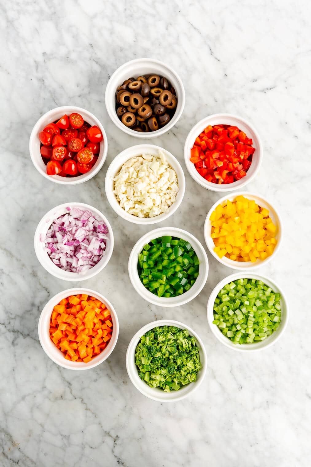 All of the chopped veggie ingredients for an Italian pasta salad in separate little white bowls on a marble surface.