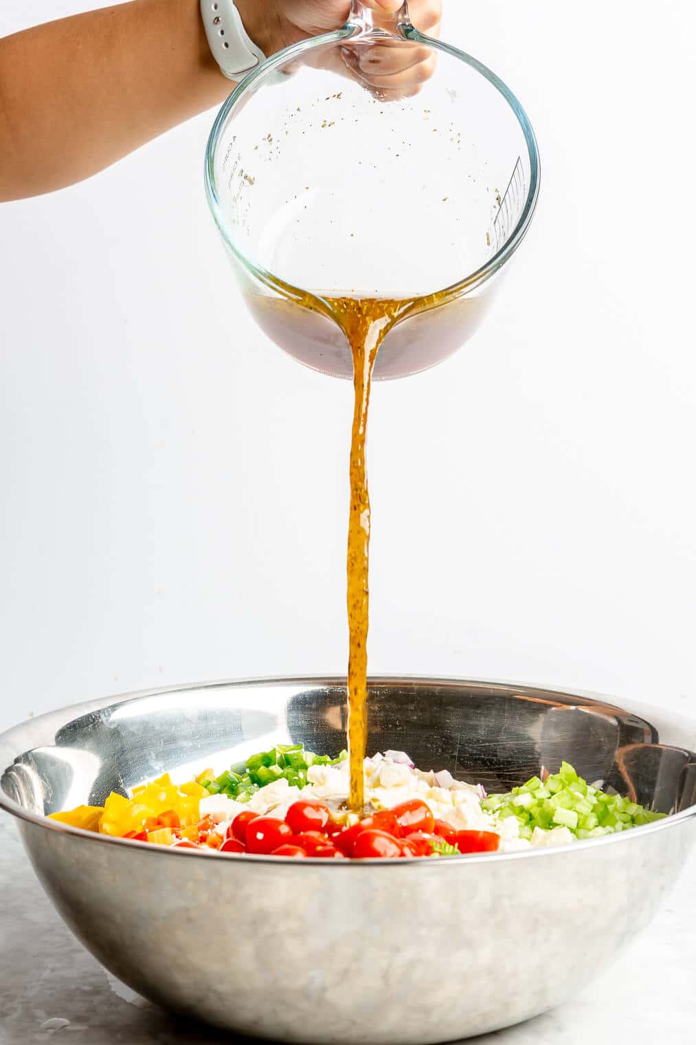 A person pouring homemade dressing into a large metal bowl to make Italian pasta salad.