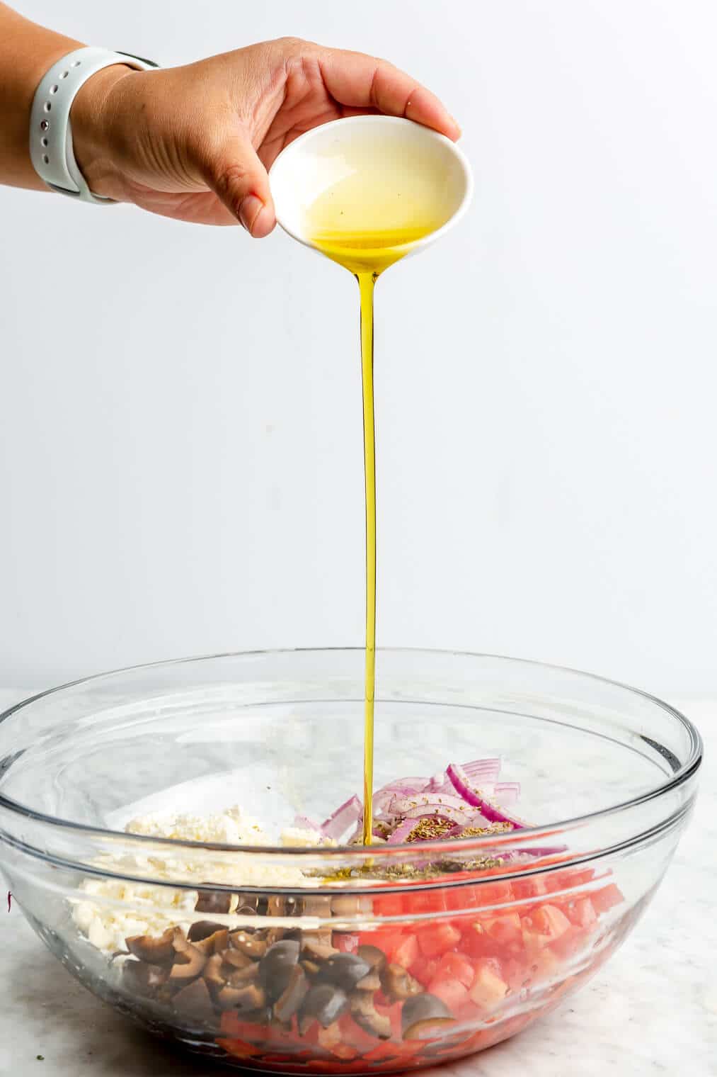 A person adding olive oil into a large glass bowl of other ingredients for Mediterranean hummus wraps.