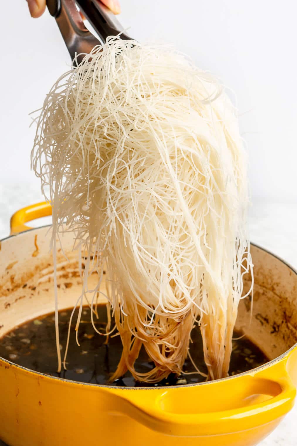 A person adding rice noodles into a large pot of sauce to make pancit.