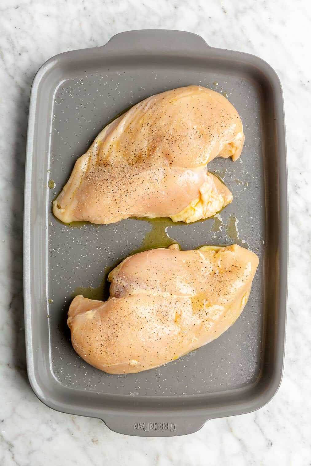 Two raw chicken breasts seasoned with salt, pepper, and olive oil on a sheet pan.
