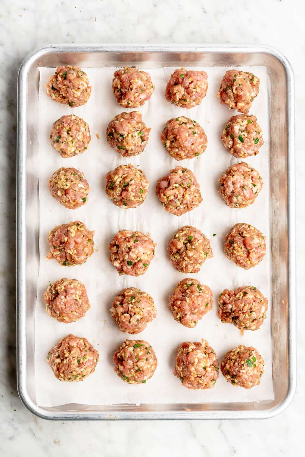24 Swedish meatballs (raw) lined up on a parchment paper-lined sheet pan before going into the oven.