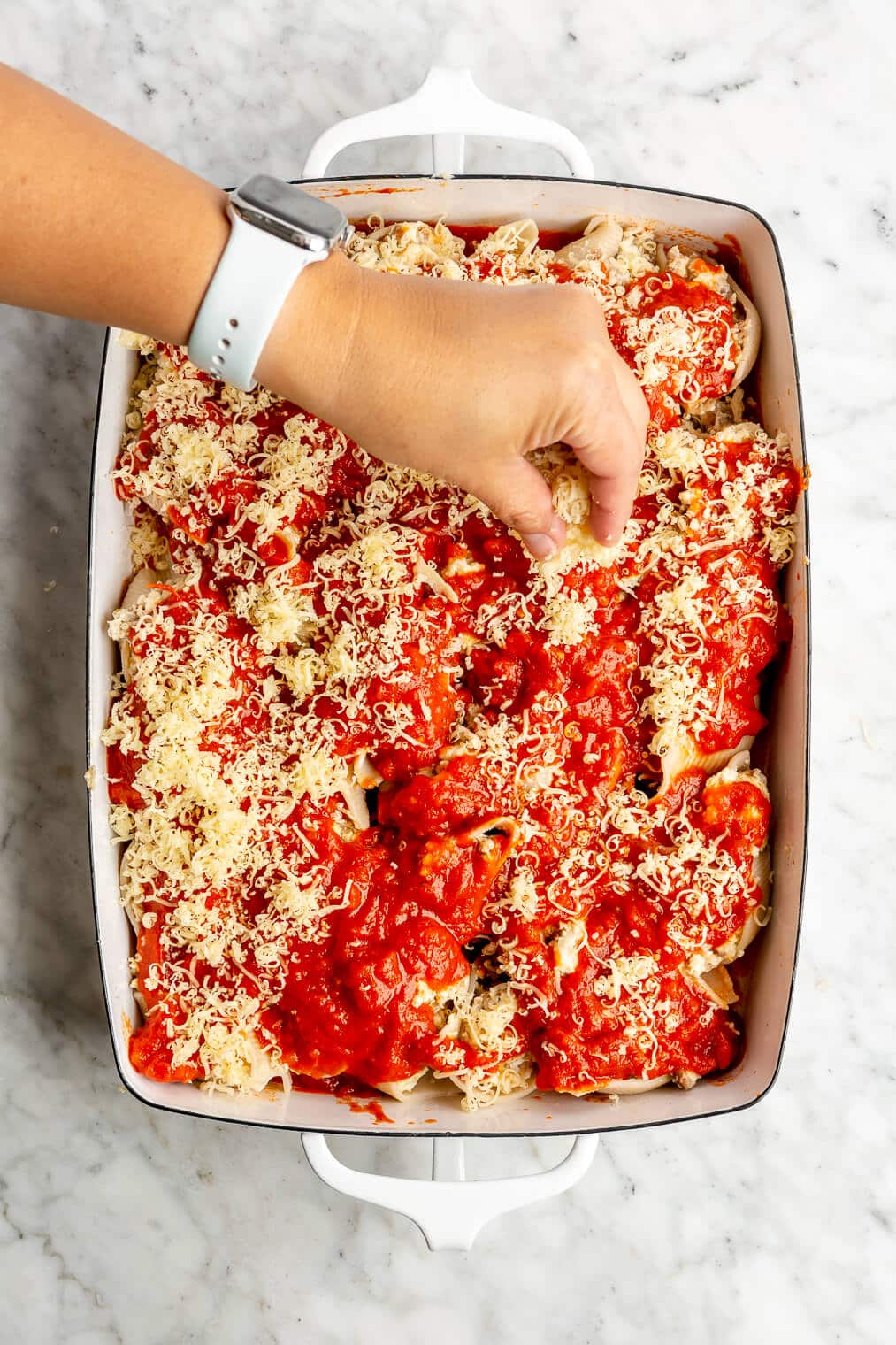 A person sprinkling shredded mozzarella cheese over top stuffed pasta shells topped with marinara sauce.