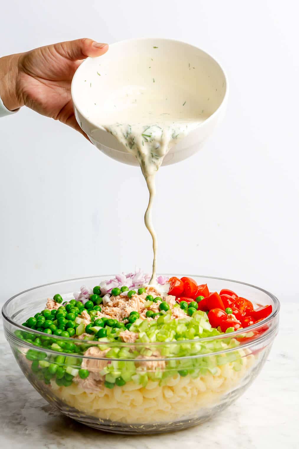 A person pouring a creamy pasta salad dressing into a large glass bowl with all of the other tuna pasta salad ingredients.