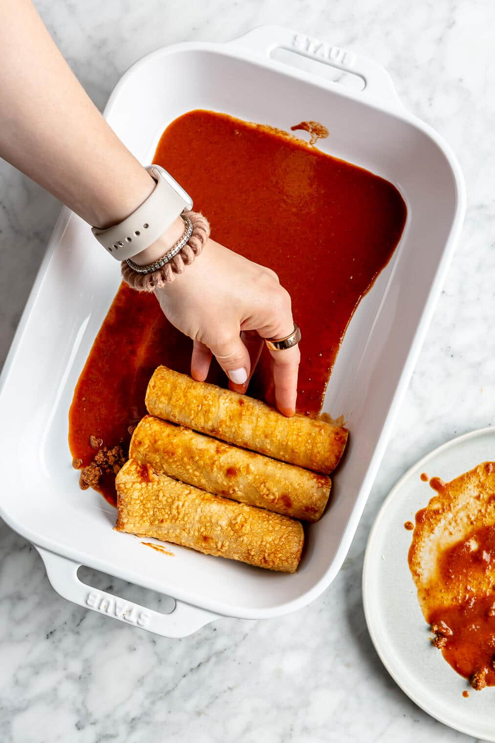 A person placing a rolled enchilada into a casserole dish next to two other enchiladas.