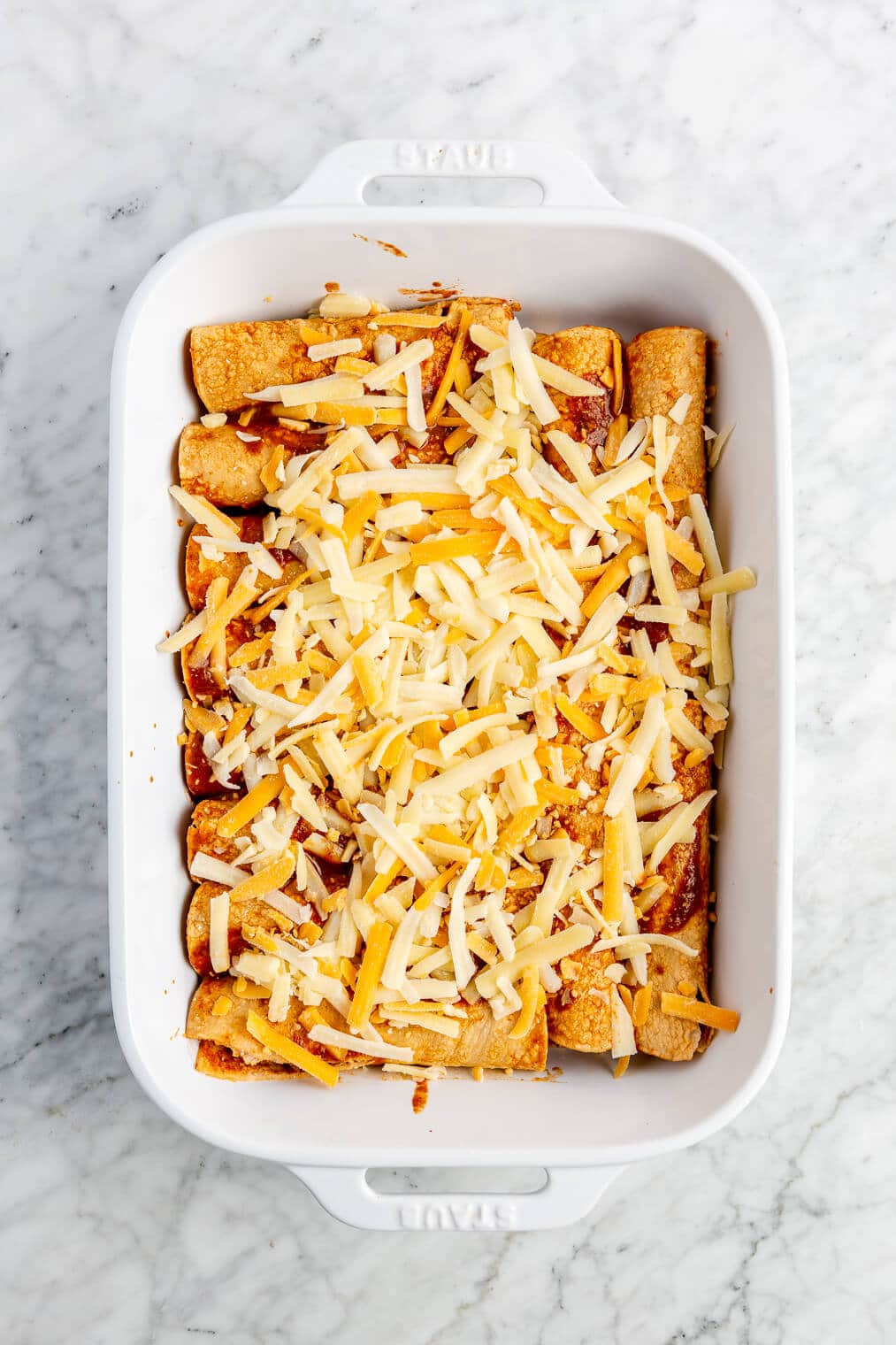 12 enchiladas topped with enchilada sauce and cheese lined up in a casserole dish before going into the oven.