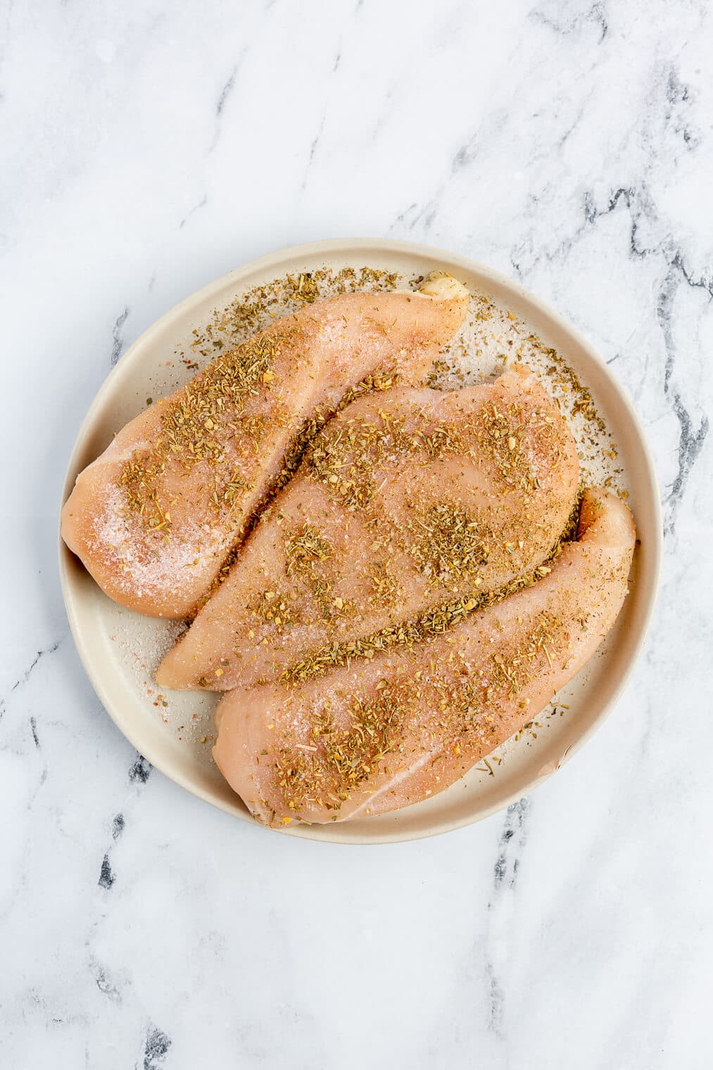 3 raw chicken breasts dusted with salt and Italian seasoning sitting on a plate.