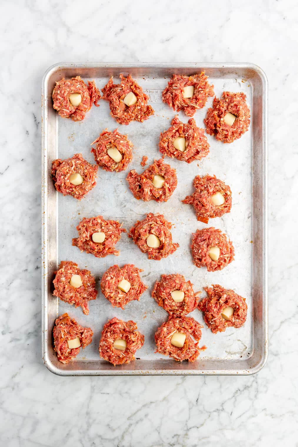 Raw pizza meatballs with a piece of a cheese stick in the middle of each one on a sheet pan before being rolled.