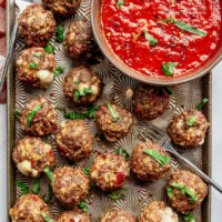 Pizza meatballs on a decorative sheet pan next to a bowl of pizza sauce. Everything is topped with fresh basil and red pepper flakes.