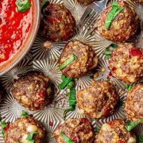 Pizza meatballs on a decorative sheet pan next to a bowl of pizza sauce. Everything is topped with fresh basil and red pepper flakes.
