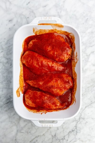 4 raw chicken breasts covered in BBQ sauce and spices in a white baking dish.