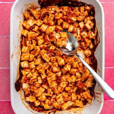 Baked BBQ Chicken Breasts cubed in a large white baking dish on a hot pink tiled background.