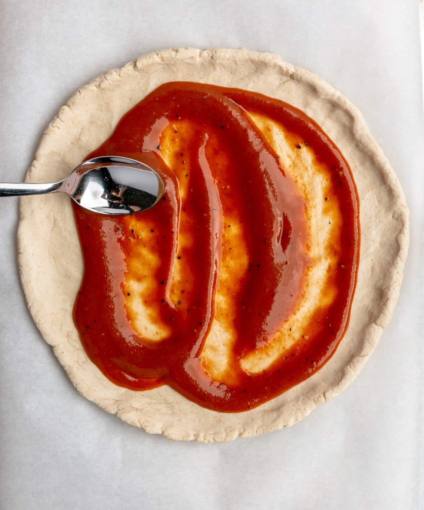 A person using a spoon to spread BBQ sauce over pizza dough.