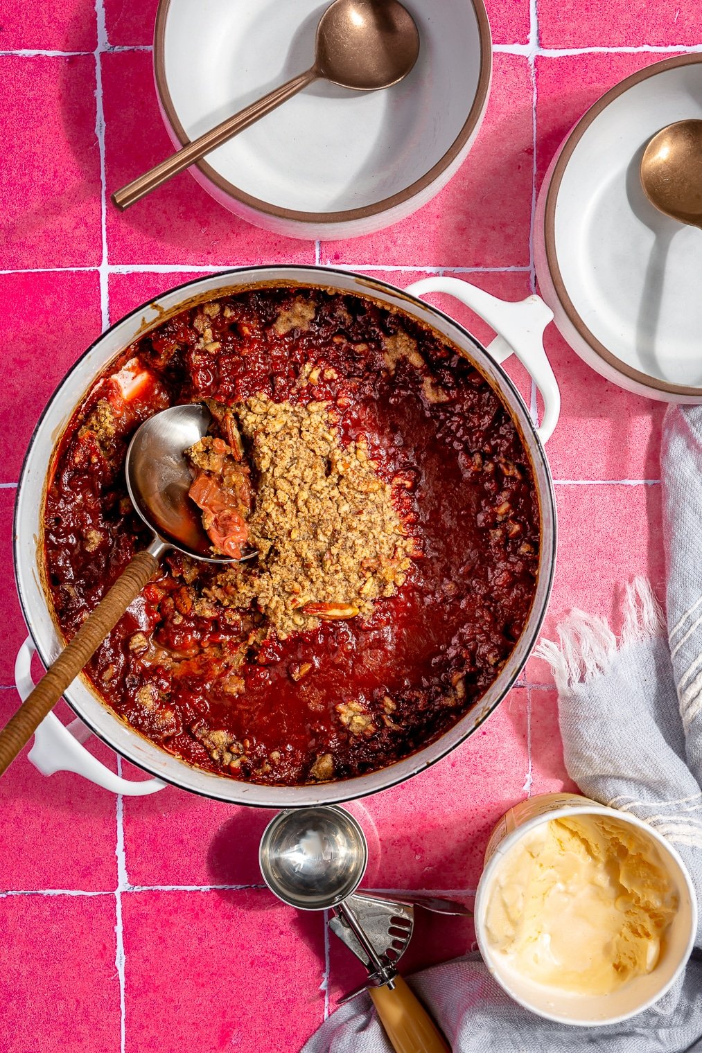 A large dish of strawberry rhubarb crisp sitting on a pink tiled background next to two empty bowls and a small carton of vanilla ice cream.