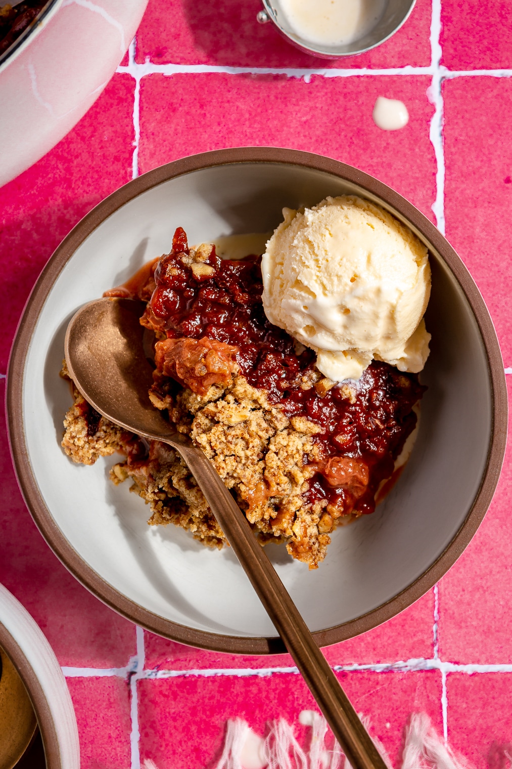 A bowl of strawberry rhubarb crisp topped with a scoop of vanilla ice cream sitting on a pink tiled background.