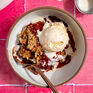 A bowl of strawberry rhubarb crisp topped with a scoop of vanilla ice cream sitting on a pink tiled background.