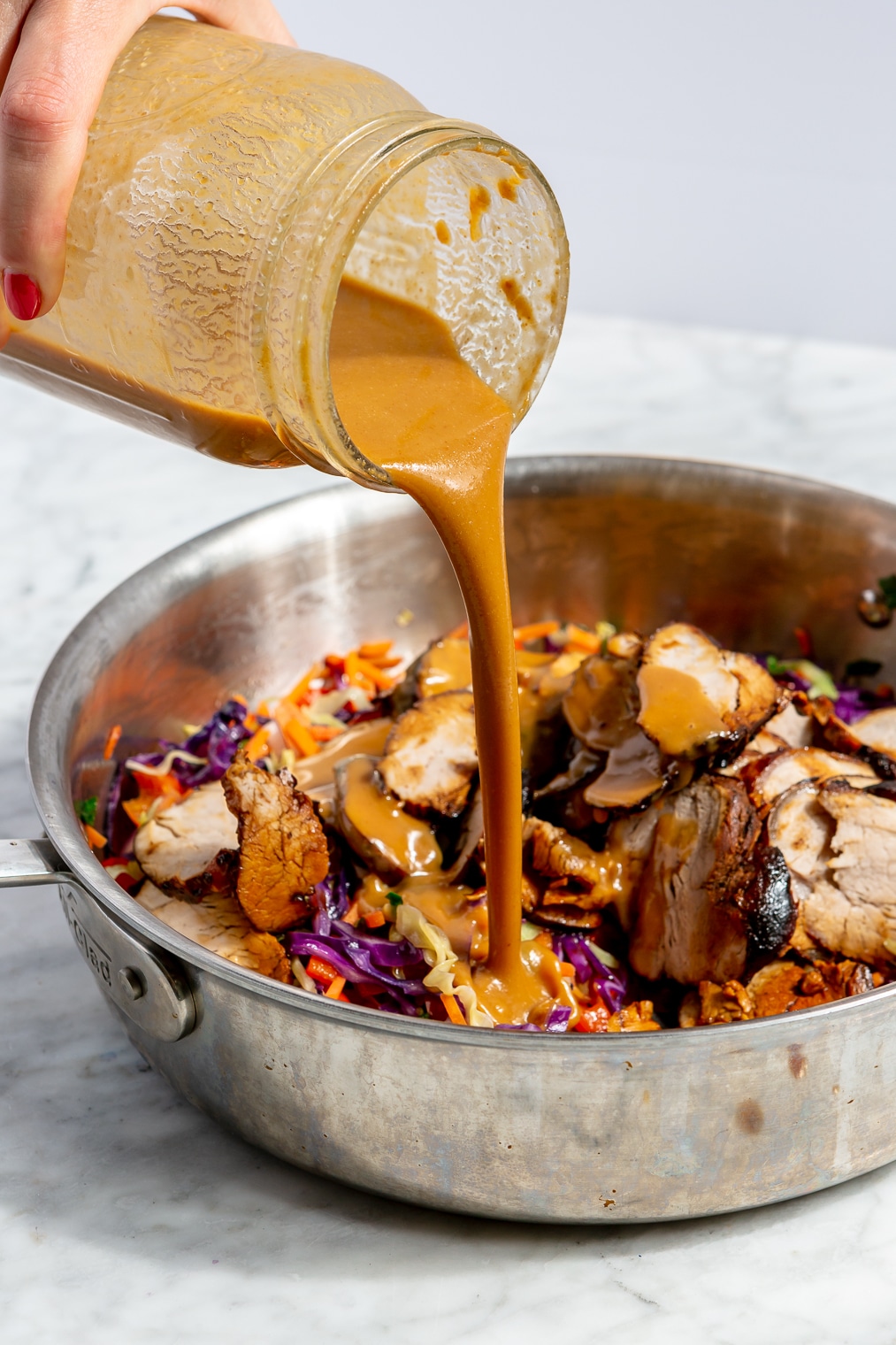 A person pouring peanut teriyaki sauce over a pork and veggie stir fry in a pan.