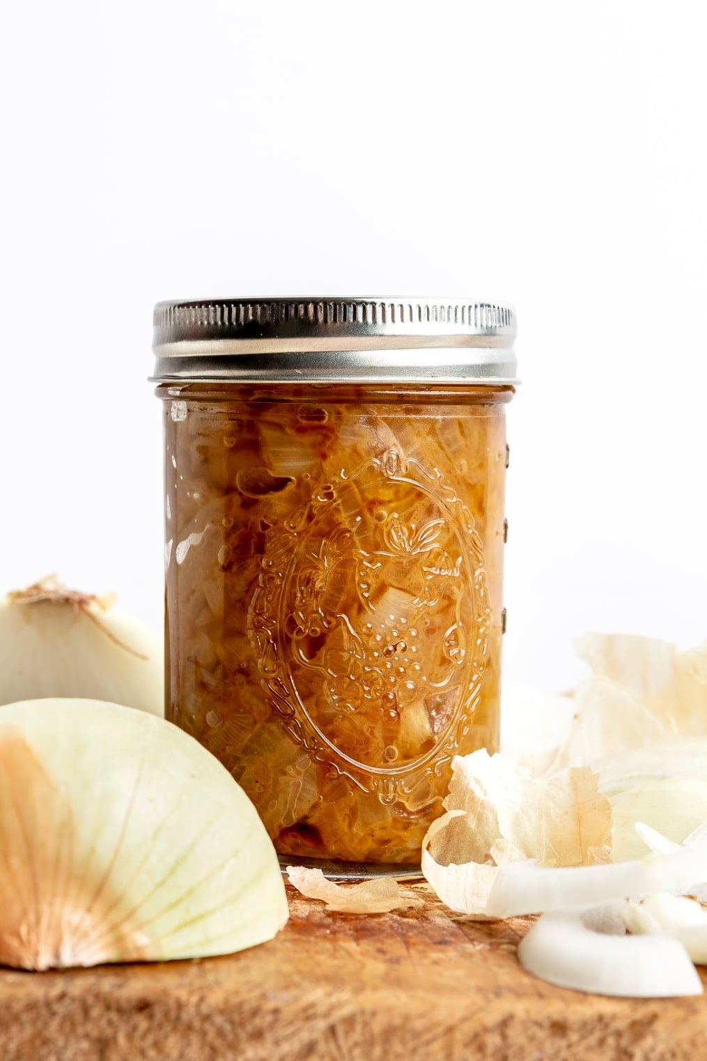 A small mason jar full of caramelized onions sitting on a wooden cutting board next to sliced and whole onions.