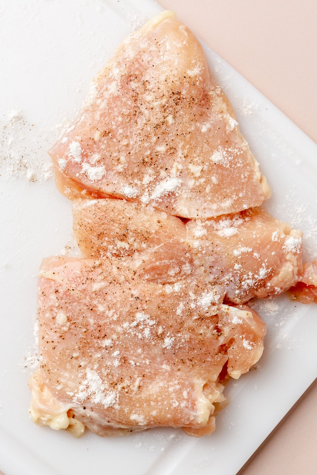 Two raw chicken breasts dusted with flour, salt, and pepper on a cutting board.