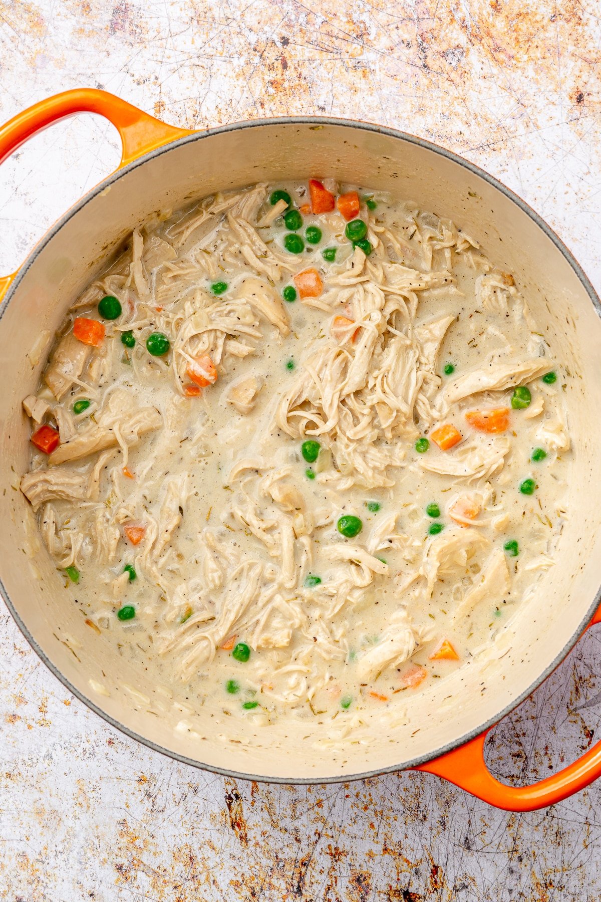 A creamy mixture with shredded chicken, diced carrots, and peas sit in an enameled pot.