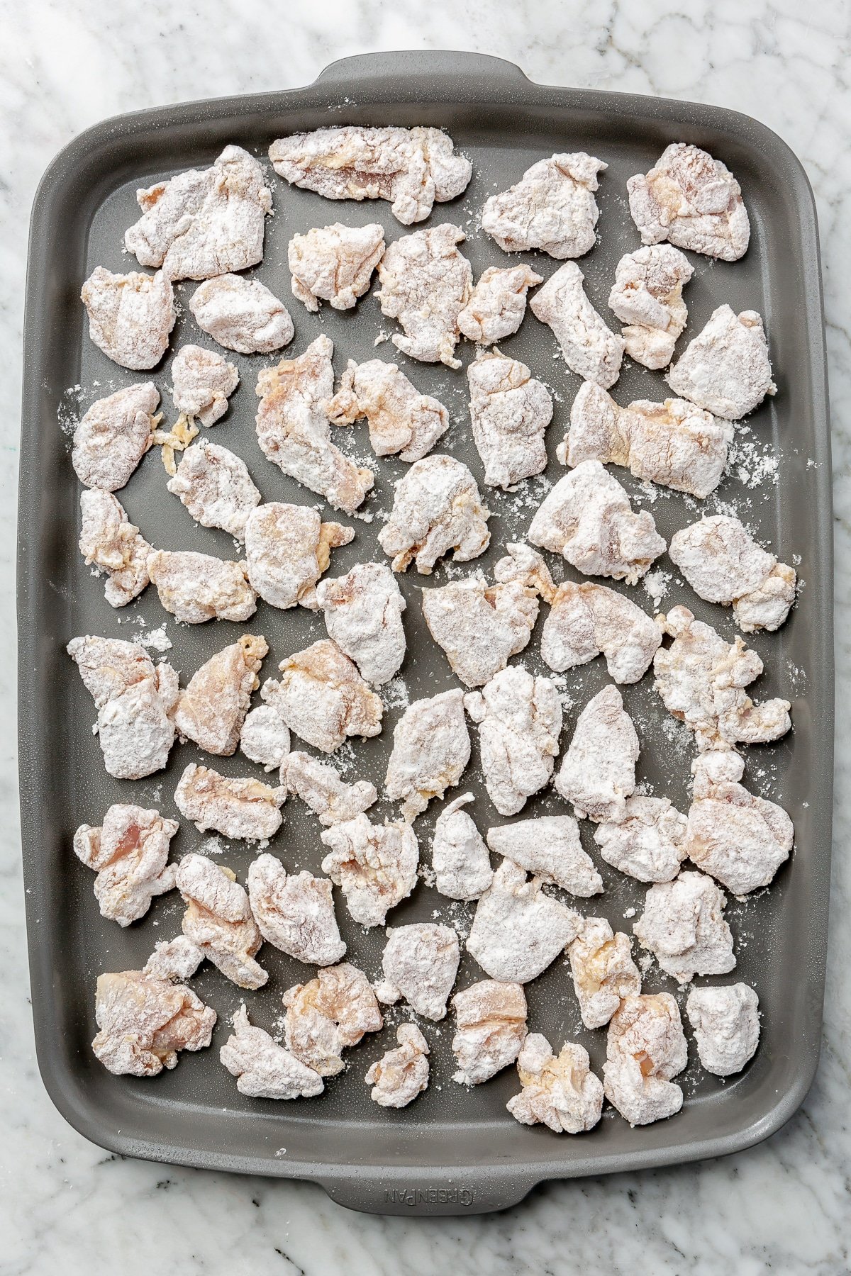 Floured chicken cubes lay, evenly distributed, on a nonstick cooking pan.