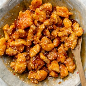 Battered, baked chicken pieces is tossed in a red sauce. White sesame seeds are sprinkled on top. A wooden spoon is placed in the same bowl.