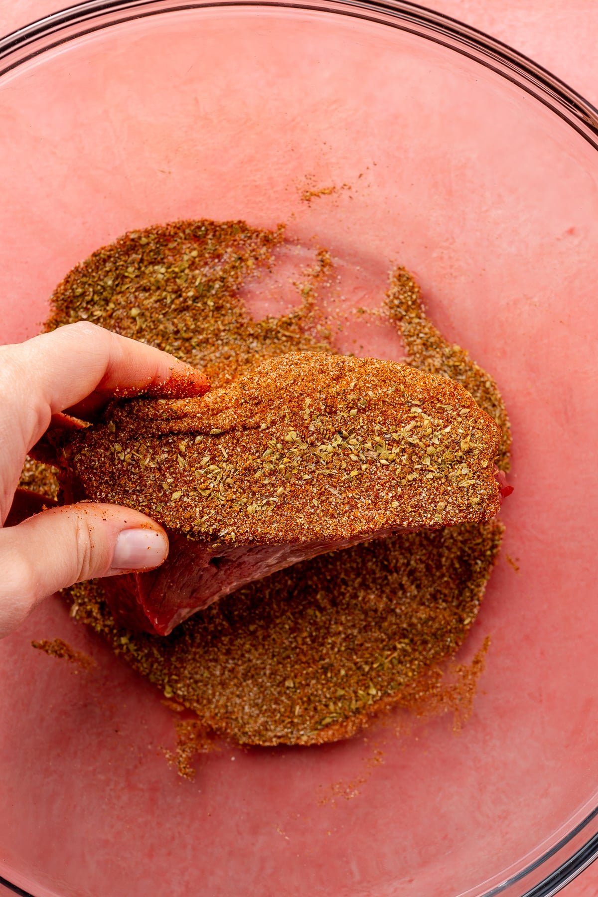 A hand is shown rolling a rectangular piece of beef brisket in a glass bowl of mixed spices.