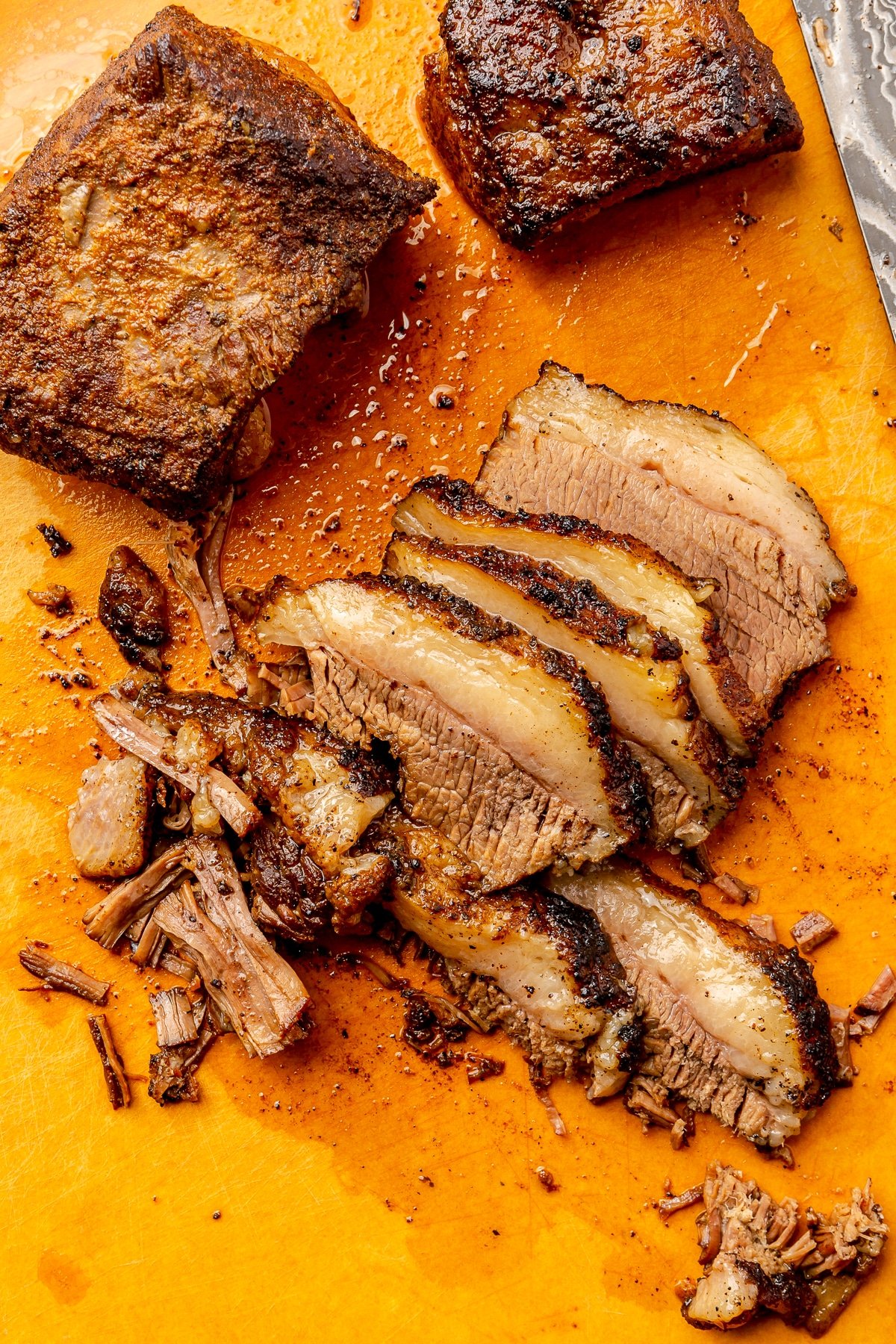 Fully cooked beef brisket has been sliced and laid out on an orange cutting board. Two, uncut pieces sit in the back.