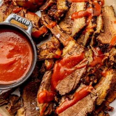 Sliced beef brisket sits on a brown plate. It has been drizzled with a red barbecue sauce. A bowl of sauce sits on the side.