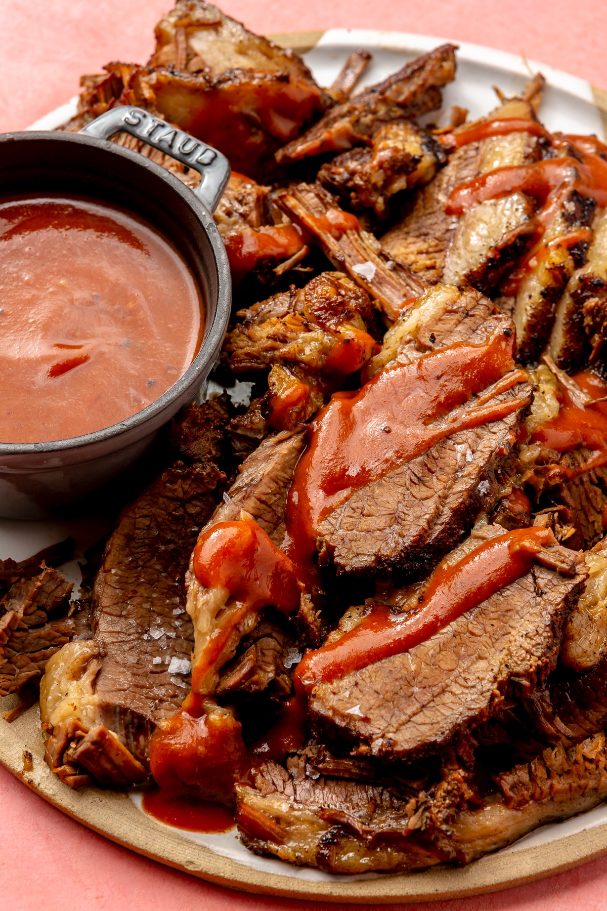 Sliced beef brisket sits on a brown plate. It has been drizzled with a red barbecue sauce. A bowl of sauce sits on the side.