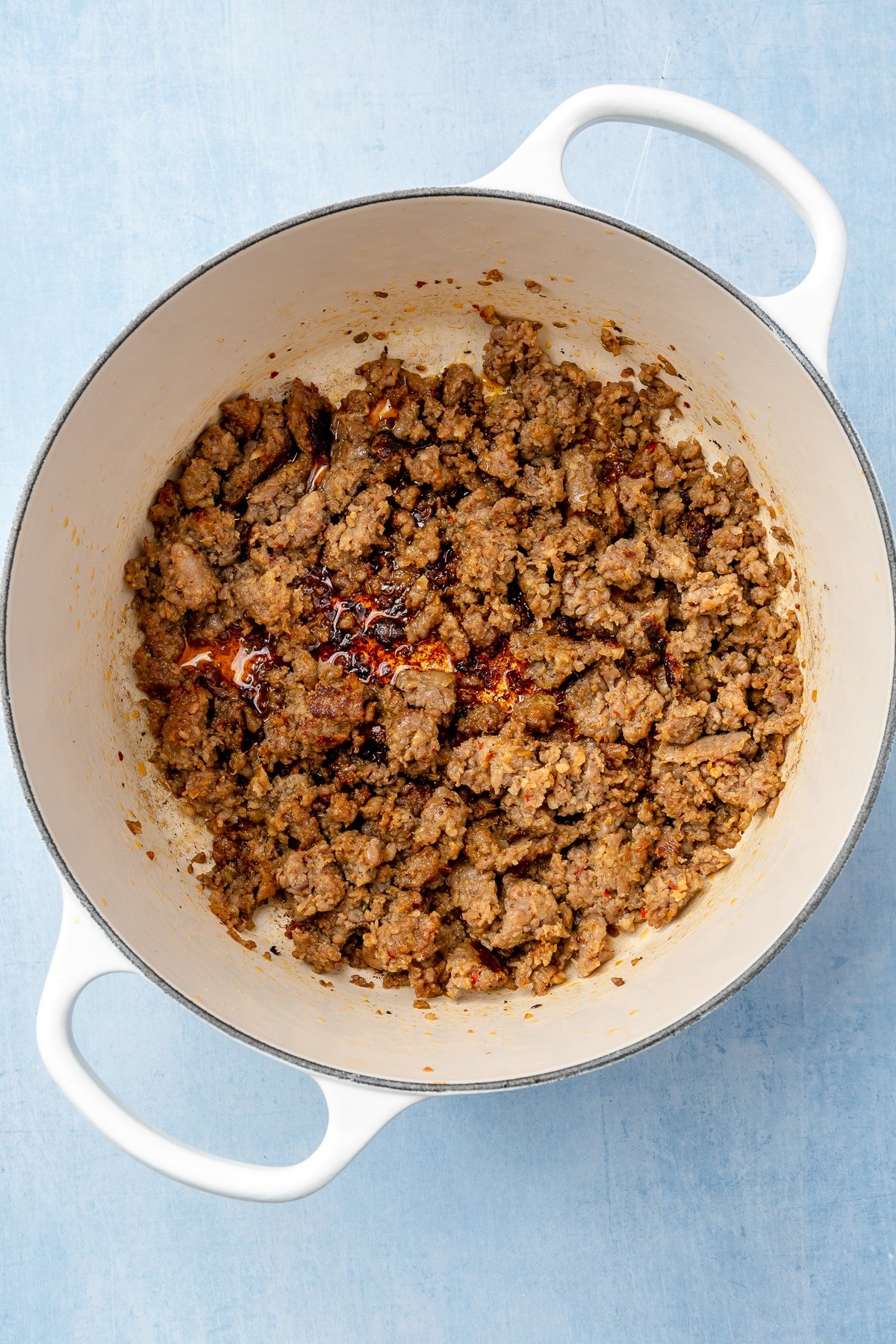 Sausage has been crumbled and cooked to a light brown in a white, enameled pot.