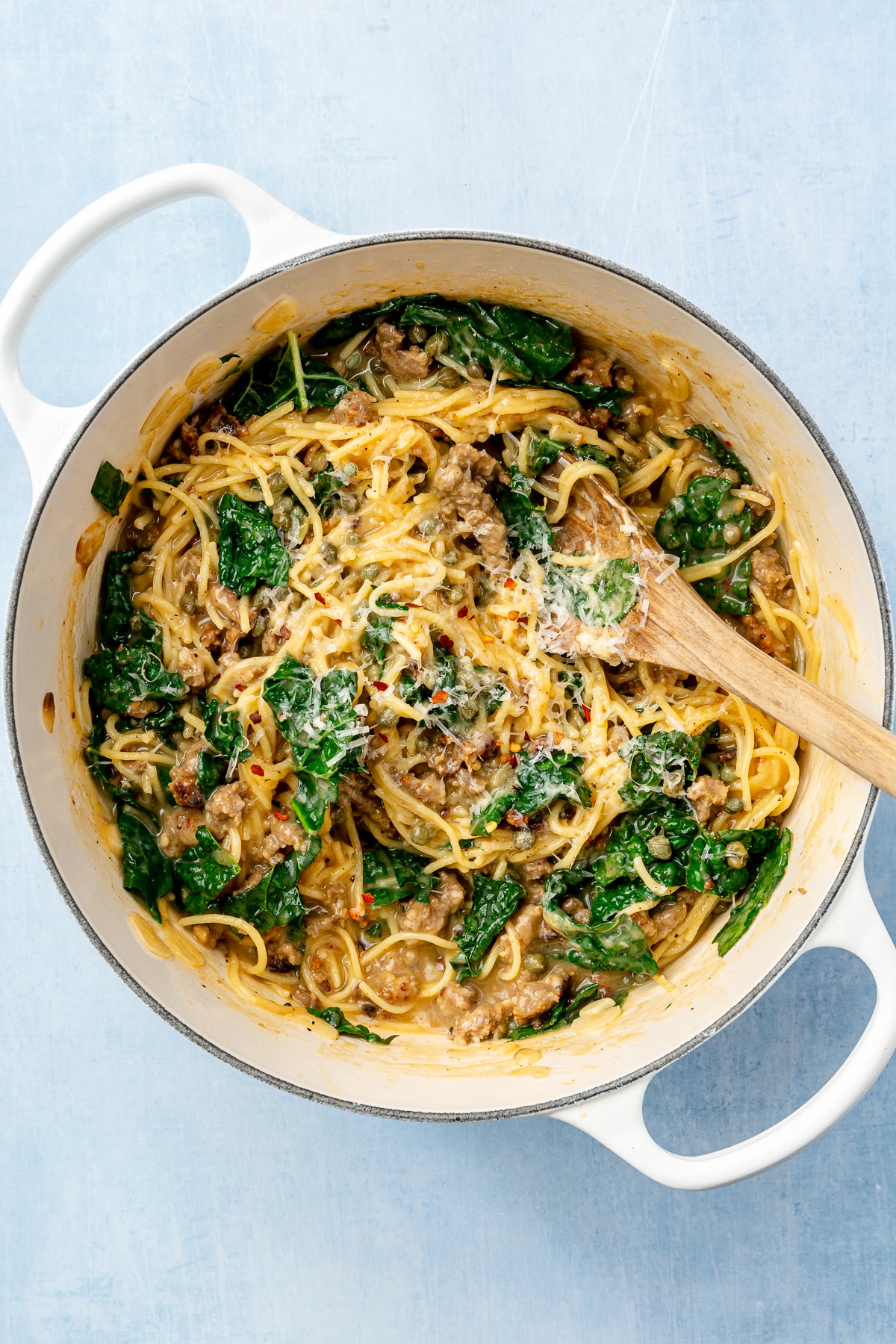 Parmesan cheese has been stirred into the kale, sausage, and spaghetti mixture. The wooden spoon, used to stir, sits in the pot.