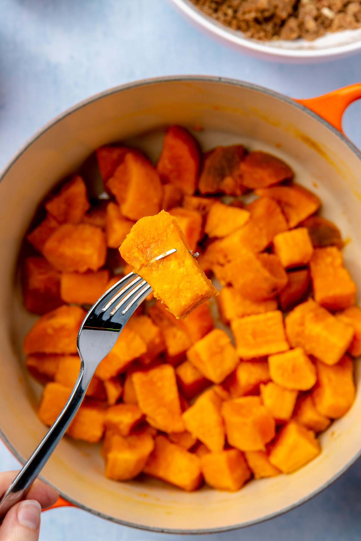 A piece of softened sweet potato is held with a fork over an orange, enameled pot of more softened, cubed sweet potato.