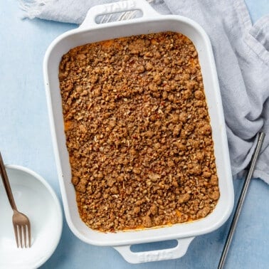 A baked sweet potato mixture, topped with a brown crumble sits, baked, in a white baking dish. A grey dish towel and a large serving spoon sit to the right-hand side. Two white bowls, each with a fork, sit to the left.