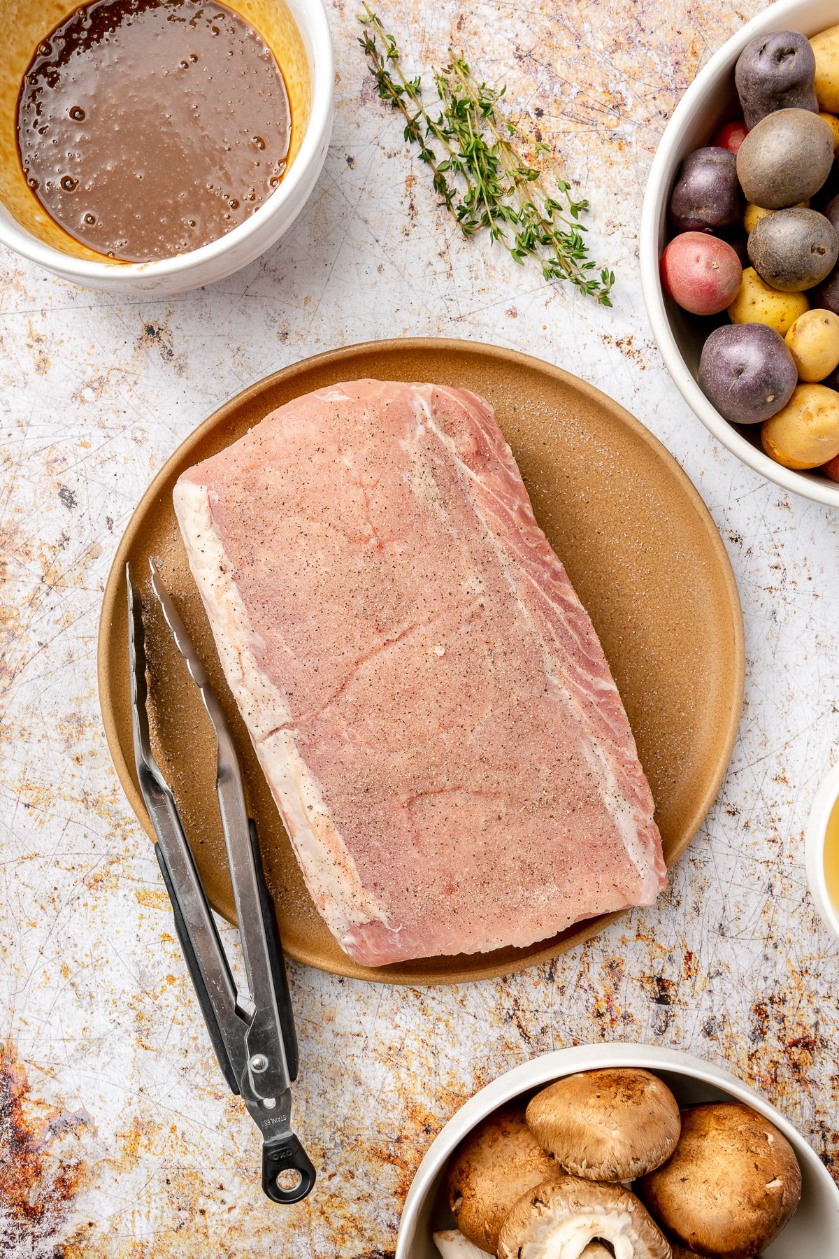 The seasoning mixture has been rubbed on the surface of a pork loin which sits on a brown plate. A black set of tongs sit on the side.