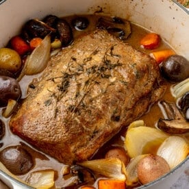 Fully cooked balsamic pork loin roast sits in a white, enameled pot. Two glasses of wine, a brown plate with golden silverware, and a chopping block sit on the side.