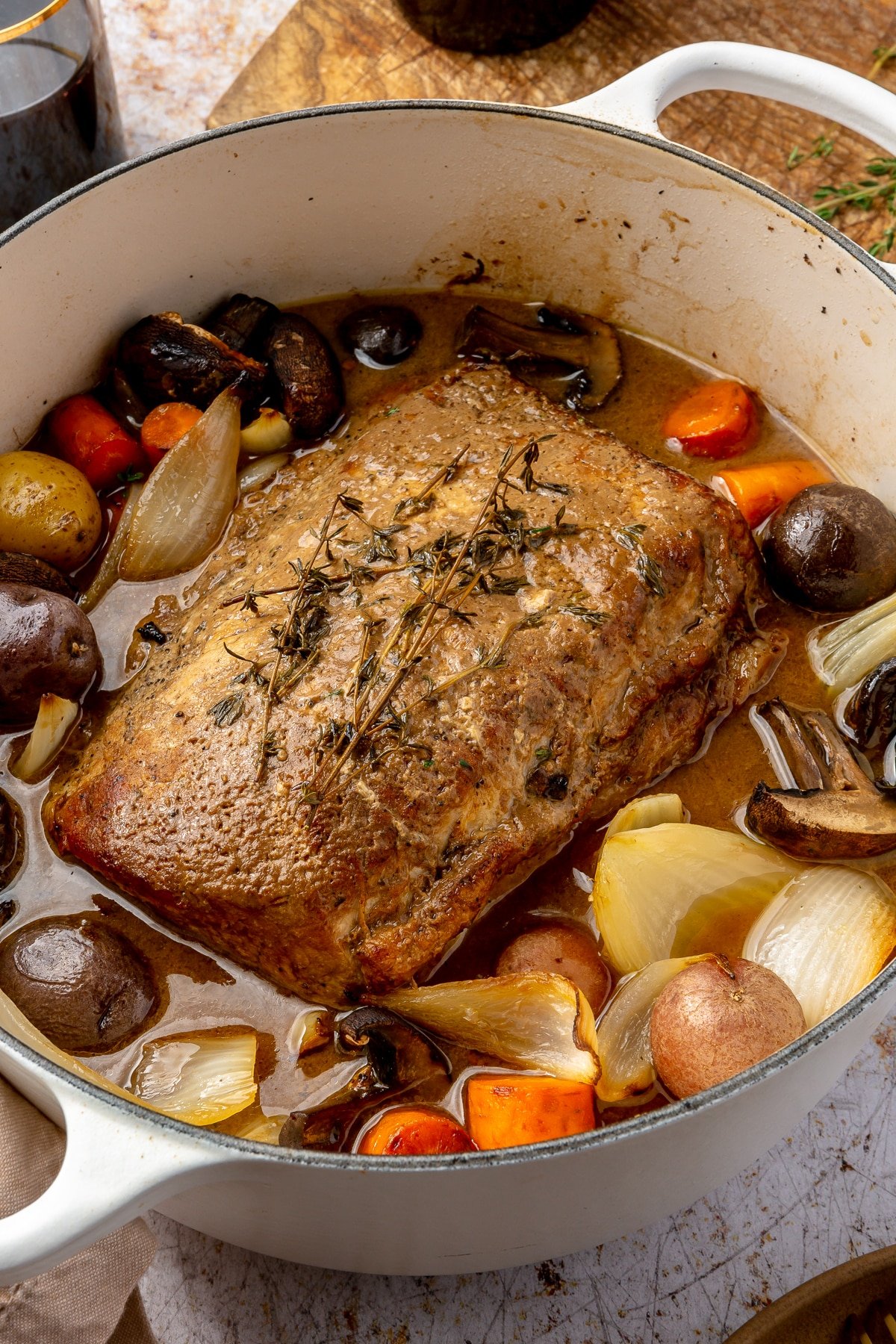 Fully cooked balsamic pork loin roast sits in a white, enameled pot. Two glasses of wine, a brown plate with golden silverware, and a chopping block sit on the side.