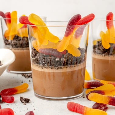 A clear glass cup layered with chocolate pudding, crushed up chocolate sandwich cookies, and gummy worms.