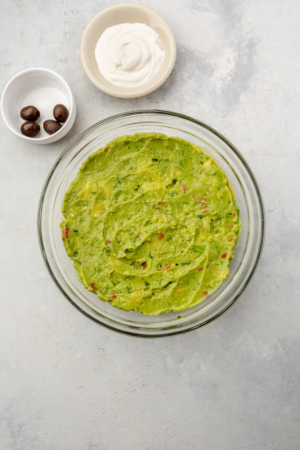 A large bowl of layered hummus, shredded cheese, pico de gallo, and guacamole (for 7 layer dip) surrounded by smaller bowls of black olives and sour cream.