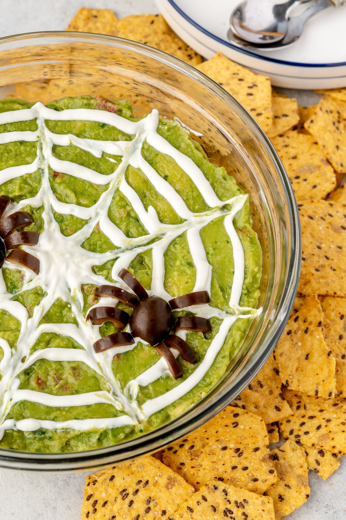 A finished bowl of spooky 7 layer dip complete with a sour cream spider web and black olive spiders on top next to corn chips.