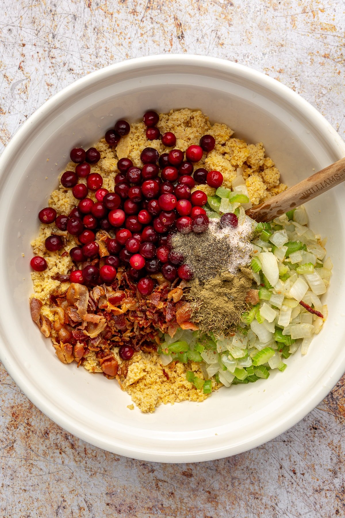 Cranberries, chopped bacon, and a variety of spices have been added to the large, white mixing bowl.