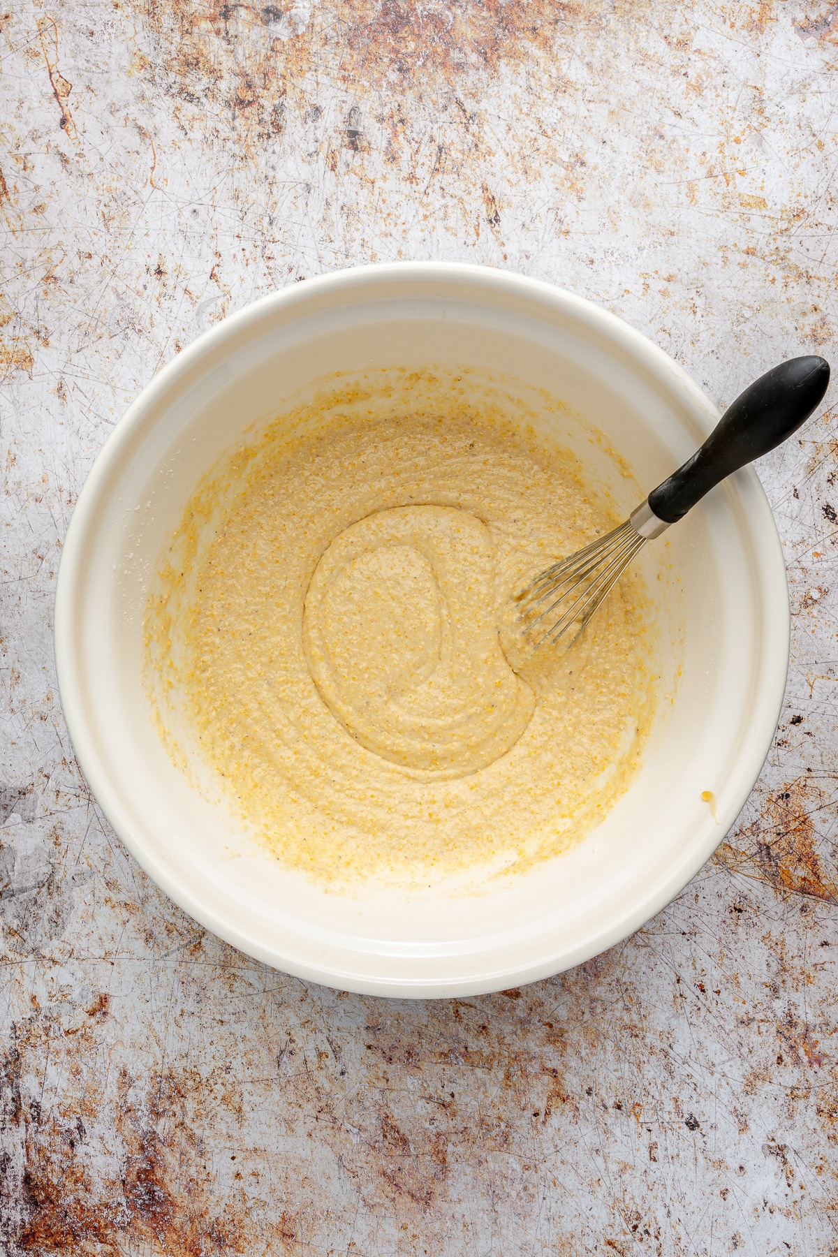 A large white bowl of cornbread batter with a whisk leaned up on the side of the bowl.