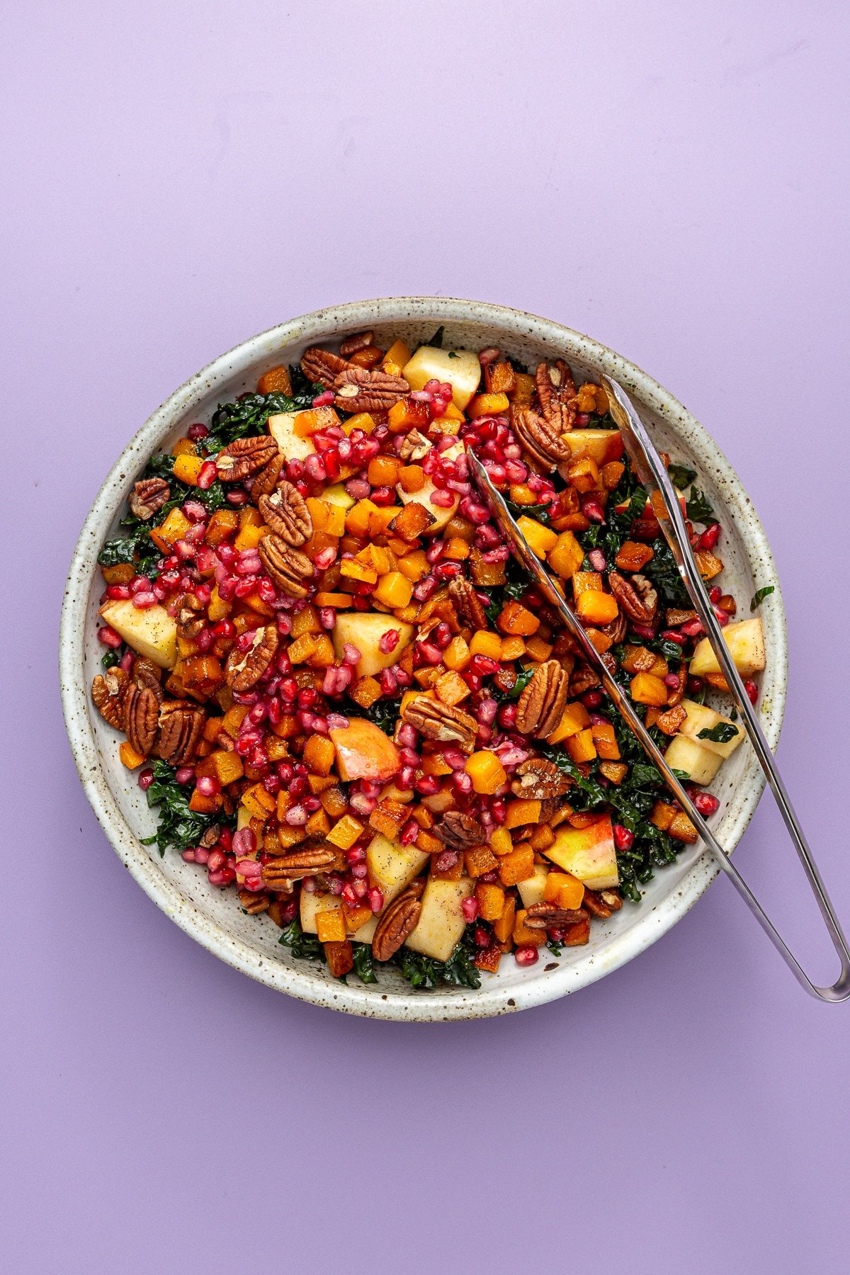 Remaining ingredients, including pomegranate seeds, pecans, and apple chunks, have been poured on top of the chopped kale. Tongs sit to the side.