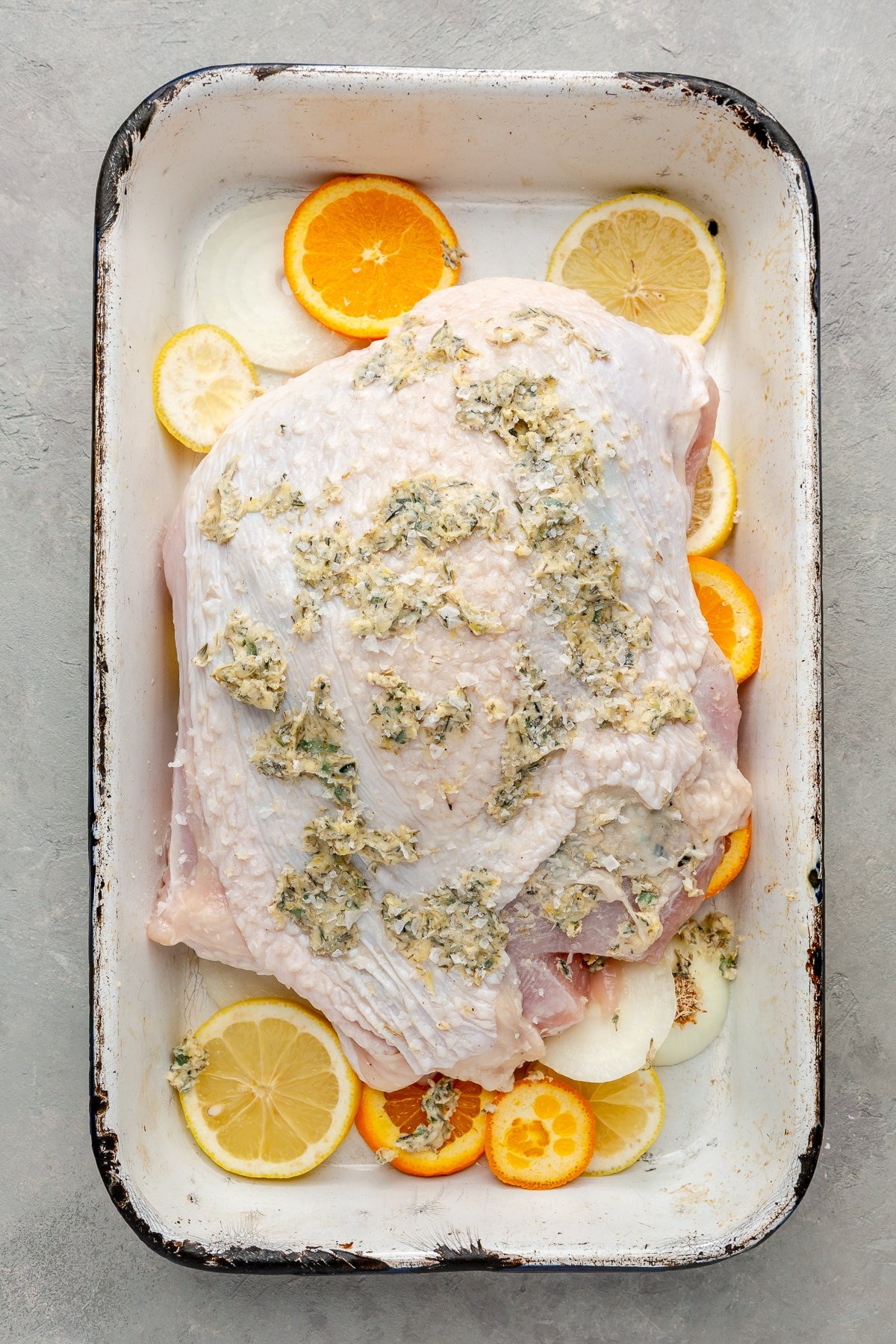 Raw turkey breast has been placed on top of the lemons, oranges, and onions. A variety of herbs and spices have been placed on top.