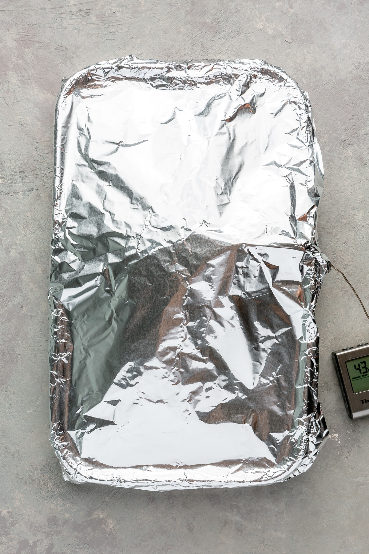 The turkey breast other ingredients in the baking pan have been covered with aluminum foil. The meat thermometer sticks out from the side.