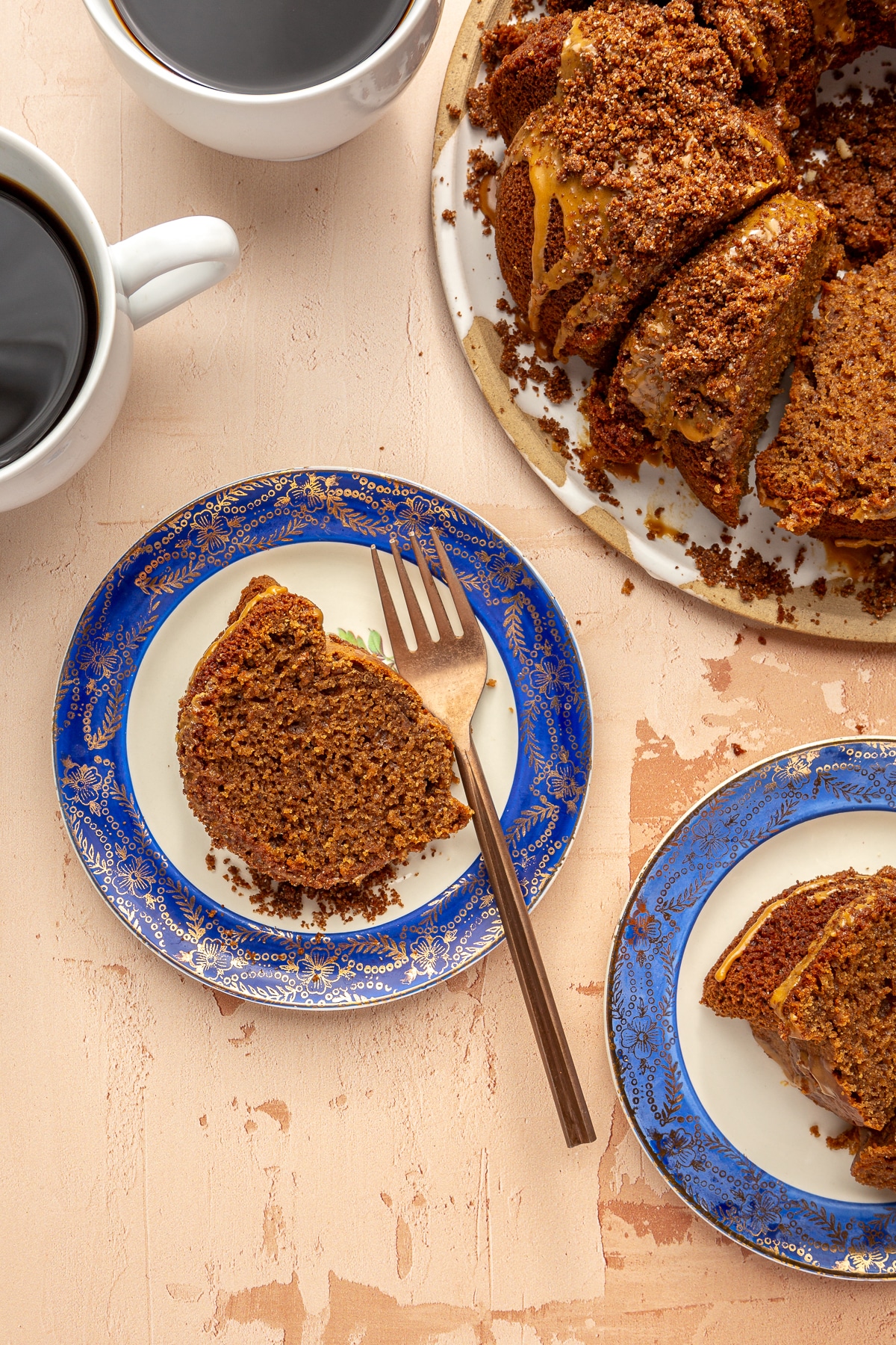 Two slices of pumpkin spice bundt cake on small blue and gold rimmed dessert plates next to two mugs of coffee and the rest of the bundt cake on a large plate.