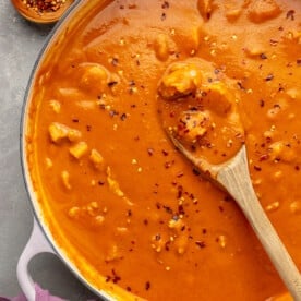 Red pepper flakes have been added to the top of the curry in the white pot. A wooden spoon is shown scooping a bit of the curry.