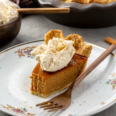 A bite has been taken out of a slice of pumpkin pie, topped with whipped cream and cinnamon. It sits on a white plate with a copper fork. The remaining pie and whipped cream sits in the background.